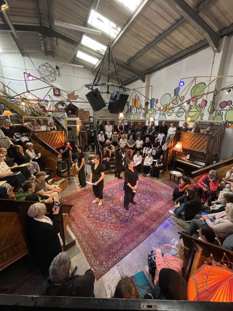 Five poetry choir members dressed in black stand in the centre of a room surrounded by an audience on raised seating
